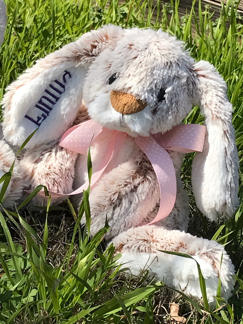 Op de afbeelding: Een pluche konijn met een roze strik en de naam "Linnea" geborduurd op &eacute;&eacute;n oor. Het konijn heeft een zachte, donzige textuur met bruine en witte vacht. Het zit in groen gras.