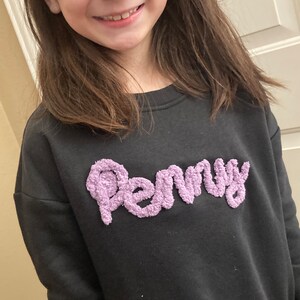 K&ouml;nnte beinhalten: Ein schwarzes Langarm-Sweatshirt mit dem Namen Penny in erhabener, strukturierter, hellvioletter Chenille-Schrift auf der Brust. Das Kleidungsst&uuml;ck hat einen Rundhalsausschnitt und ein schlichtes, einfarbiges Design mit weicher Oberfl&auml;che.