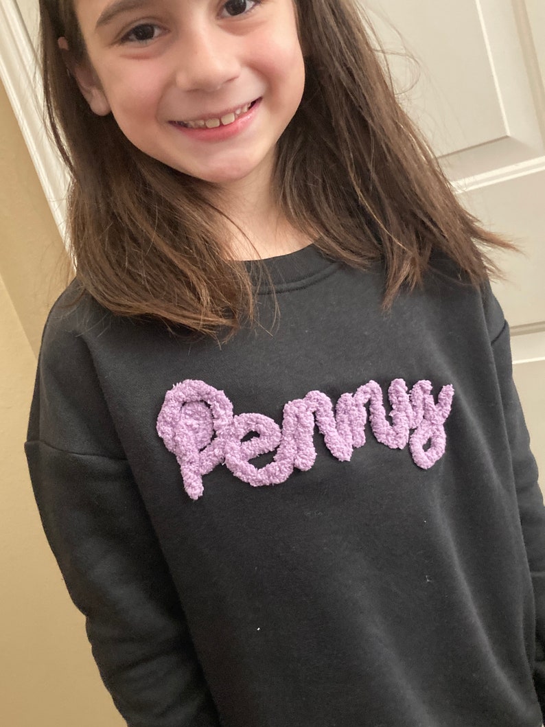K&ouml;nnte beinhalten: Ein schwarzes Langarm-Sweatshirt mit dem Namen Penny in erhabener, strukturierter, hellvioletter Chenille-Schrift auf der Brust. Das Kleidungsst&uuml;ck hat einen Rundhalsausschnitt und ein schlichtes, einfarbiges Design mit weicher Oberfl&auml;che.