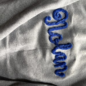 K&ouml;nnte beinhalten: Ein graumeliertes Sweatshirt mit dem vertikal gestickten Wort "Graham" aus dickem, strukturiertem, k&ouml;nigsblauem Chenillegarn. Der Stoff des Sweatshirts hat ein weiches, leicht zerknittertes Aussehen mit einer deutlich sichtbaren Strickstruktur.