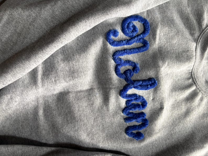 K&ouml;nnte beinhalten: Ein graumeliertes Sweatshirt mit dem vertikal gestickten Wort "Graham" aus dickem, strukturiertem, k&ouml;nigsblauem Chenillegarn. Der Stoff des Sweatshirts hat ein weiches, leicht zerknittertes Aussehen mit einer deutlich sichtbaren Strickstruktur.
