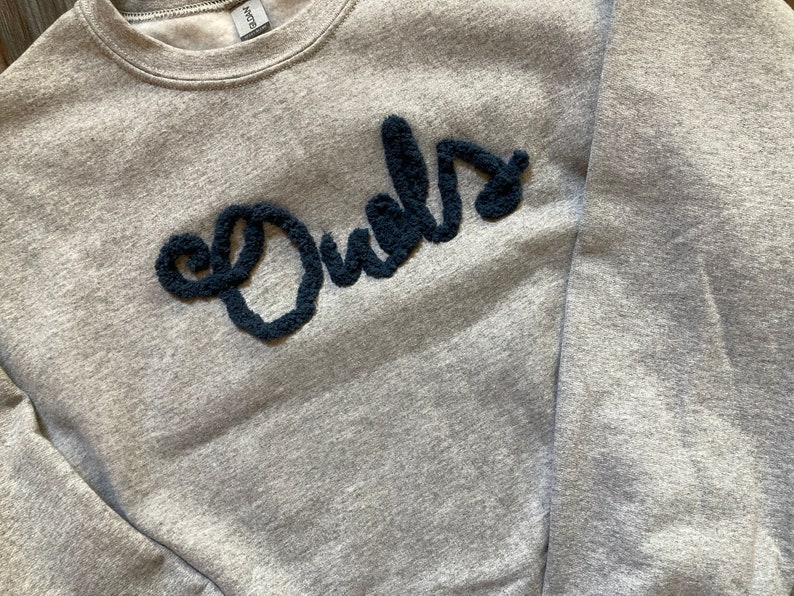 K&ouml;nnte beinhalten: Ein graumeliertes Sweatshirt mit Rundhalsausschnitt und dem Wort Owls in dunkelblauer, strukturierter, flauschiger und erhabener Garnschrift auf der Brust. Das Kleidungsst&uuml;ck hat lange &Auml;rmel, einen gerippten Kragen und ein Fleecefutter.