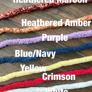 K&ouml;nnte beinhalten: Sieben Str&auml;nge aus flauschigem Garn, horizontal angeordnet in den Farben Heathered Maroon, Heathered Amber, Purple, Blue/Navy, Yellow, Crimson und White. Jeder Strang hat eine weiche, strukturierte Optik und liegt in einer parallelen Reihe.