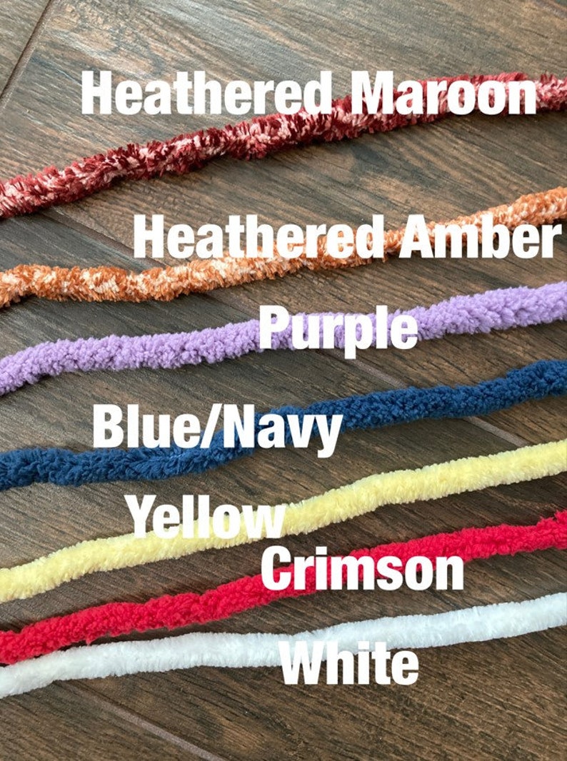 K&ouml;nnte beinhalten: Sieben Str&auml;nge aus flauschigem Garn, horizontal angeordnet in den Farben Heathered Maroon, Heathered Amber, Purple, Blue/Navy, Yellow, Crimson und White. Jeder Strang hat eine weiche, strukturierte Optik und liegt in einer parallelen Reihe.