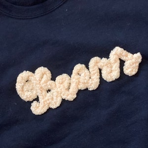 K&ouml;nnte beinhalten: Ein schwarzes Sweatshirt mit dem Wort "beam", das in cremefarbenem, strukturiertem und flauschigem Garn gestickt ist. Der Schriftzug ist in Schreibschrift gehalten und hebt sich mit einer erhabenen, weichen Optik vom dunklen Stoff ab.