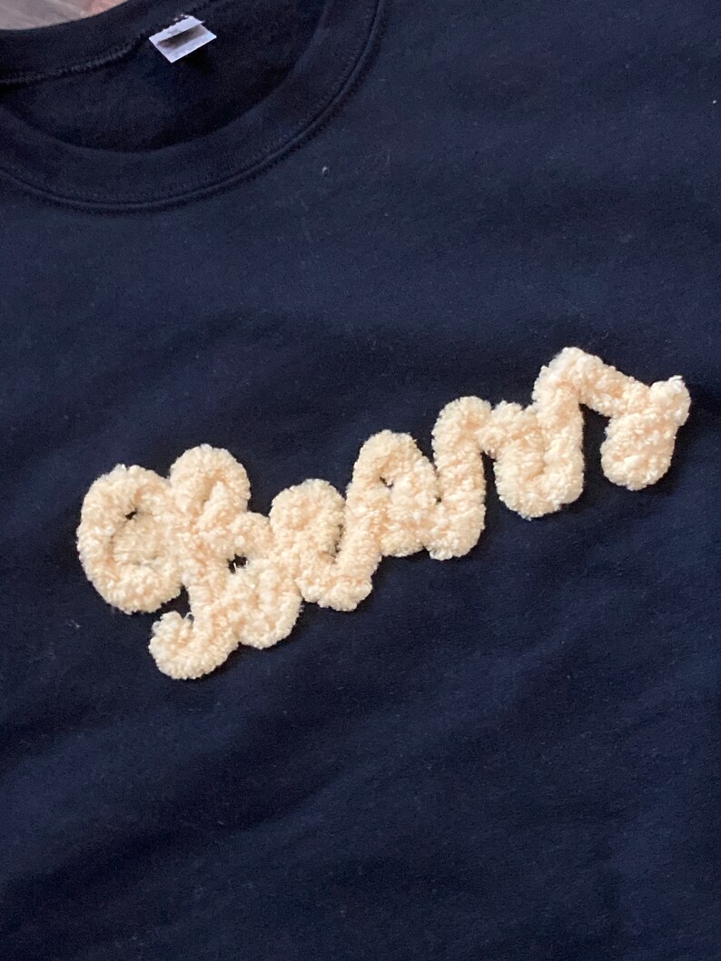 K&ouml;nnte beinhalten: Ein schwarzes Sweatshirt mit dem Wort "beam", das in cremefarbenem, strukturiertem und flauschigem Garn gestickt ist. Der Schriftzug ist in Schreibschrift gehalten und hebt sich mit einer erhabenen, weichen Optik vom dunklen Stoff ab.
