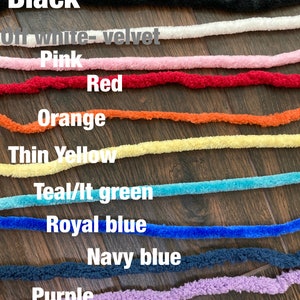 K&ouml;nnte beinhalten: Eine Sammlung von zehn flauschigen Garnstr&auml;ngen in verschiedenen Farben mit den Beschriftungen Black, Off white-velvet, Pink, Red, Orange, Thin Yellow, Teal/lt green, Royal blue, Navy blue, Purple und Off white-fluffy, horizontal gestapelt.