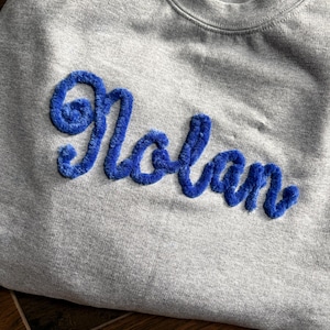 K&ouml;nnte beinhalten: Ein graumeliertes Sweatshirt mit Rundhalsausschnitt, auf dessen Brust der Name Nolan in einer leuchtend blauen, strukturierten und flauschigen Chenille-Schrift gestickt ist. Das Kleidungsst&uuml;ck hat einen gerippten Ausschnitt und ein weiches Fleecefutter.