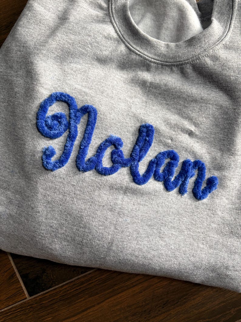 K&ouml;nnte beinhalten: Ein graumeliertes Sweatshirt mit Rundhalsausschnitt, auf dessen Brust der Name Nolan in einer leuchtend blauen, strukturierten und flauschigen Chenille-Schrift gestickt ist. Das Kleidungsst&uuml;ck hat einen gerippten Ausschnitt und ein weiches Fleecefutter.