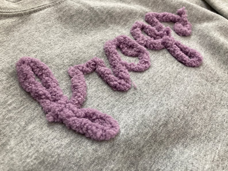 K&ouml;nnte beinhalten: Ein graumeliertes Sweatshirt mit dem Wort "knots", das in erhabenem, strukturiertem violettem Garn gestickt ist. Der Schriftzug ist in Schreibschrift gehalten, wobei der flauschige, geschlungene Flor des violetten Garns einen 3D-Effekt erzeugt.