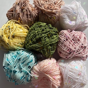 May include: A collection of nine yarn balls in various colors including beige, white, yellow, olive green, light pink, and turquoise. Each ball of yarn is interwoven with small, reflective, circular sequins that catch the light, creating a textured appearance.