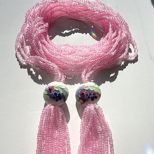 May include: A long necklace featuring multiple strands of translucent light pink glass beads. The necklace is accented by two round white glass beads, each decorated with a painted floral pattern in shades of green, blue, and red, ending in pink tassels.