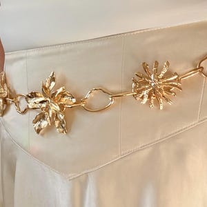 May include: A gold-toned belt with floral and starburst-shaped embellishments. The belt is made of linked metal pieces and is worn over a cream-colored garment. The floral and starburst designs add a decorative touch to the belt.