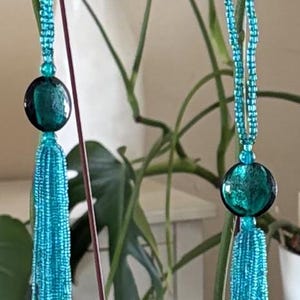May include: Two decorative hanging ornaments featuring long, vibrant turquoise beaded tassels suspended from matching turquoise beaded cords. Each ornament is accented with a single, large, round, dark teal glass bead positioned directly above the tassel.