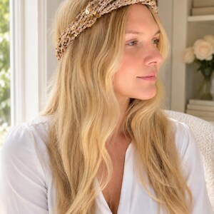 May include: A beige crochet headband featuring a twisted front design and integrated gold sequins. The accessory is crafted from a textured knit material with a subtle shimmer, providing a decorative element that sits across the top of the head.
