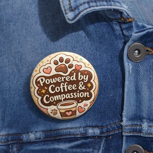May include: A round button with the text "Powered by Coffee & Compassion" in a brown and cream design. The button features paw prints, hearts, and a coffee cup illustration, set against a denim jacket background.