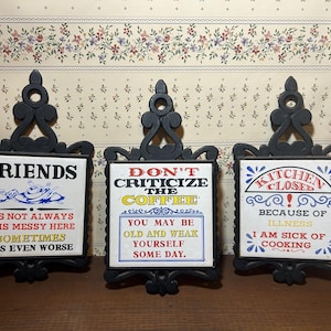Puede incluir: Tres letreros decorativos con marcos negros y citas humorísticas. Los letreros dicen "FRIENDS", "DON'T CRITICIZE THE COFFEE" y "KITCHEN CLOSED". Cada letrero tiene un fondo de azulejo blanco con texto y gráficos coloridos.