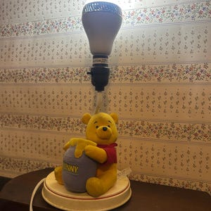 Pode incluir: Lâmpada do Ursinho Pooh com um Ursinho Pooh de pelúcia amarelo segurando um pote de mel roxo. A lâmpada tem uma base branca com uma faixa vermelha e uma lâmpada grande. O pote de mel tem a palavra "HUNNY".