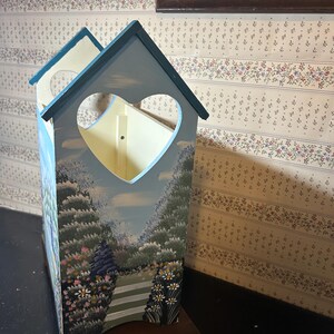 May include: A wooden decorative mailbox with a heart-shaped opening and a painted landscape scene. The mailbox is painted in shades of blue, green, and white, with a brown wooden base. The mailbox has a roof and is designed to resemble a small house.