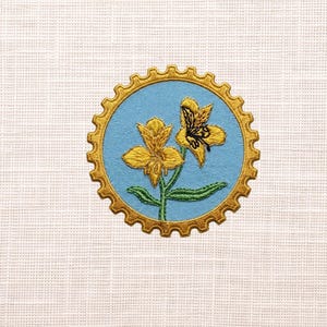 May include: Embroidered patch featuring two yellow flowers with green stems and leaves, set against a light blue background. The design is framed by a gold, gear-shaped border, all on a white fabric.