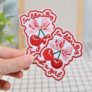 May include: Embroidered patches featuring red cherries with green stems and pink bows. The patches have the text "I'm literally just a girl" in red thread, outlined in white.