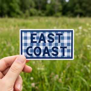 May include: A rectangular sticker with the words "EAST COAST" in navy blue block letters. The background of the sticker is a blue and white gingham pattern, with a white border. The sticker is held in front of a green grassy background.