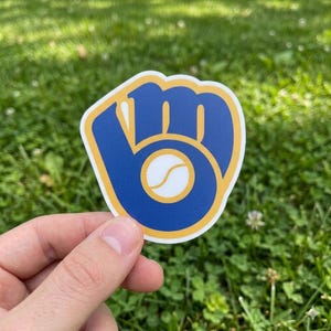 May include: A sticker featuring the Milwaukee Brewers baseball team logo. The logo is a blue baseball glove with a yellow outline, and a baseball in the center. The sticker is held against a background of green grass.