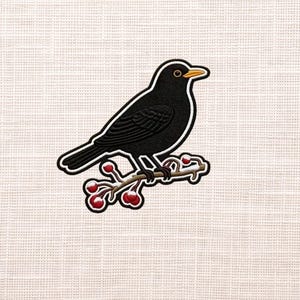 May include: A blackbird embroidered patch with a white outline, perched on a branch with red berries. The bird has a yellow beak and eye, and the patch is set against a textured, off-white background.