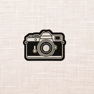 May include: A black and white embroidered patch in the shape of a vintage camera. The patch features a detailed outline of the camera body, lens, and other components. The patch is likely designed to be ironed on or sewn onto fabric.