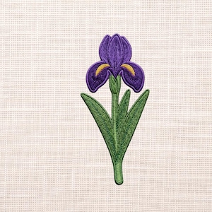 May include: An embroidered patch featuring a purple iris flower with yellow accents on the petals. The flower has a green stem and several green leaves extending downwards. The embroidery is detailed with visible thread texture throughout the design.