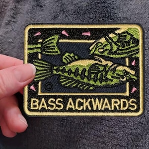 May include: Embroidered patch with a black background, a green and black fish design, and the text "BASS ACKWARDS" in gold lettering. The patch has a gold border and pink accents.
