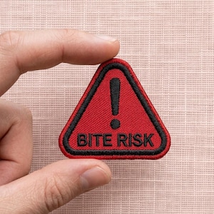 May include: A red triangular patch with a black border and an exclamation point. The text "BITE RISK" is embroidered in black. The patch is held between fingers against a textured pink background.