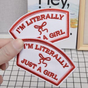 May include: Two white embroidered patches with red text and a red bow design. The text on the patches reads "I'M LITERALLY JUST A GIRL". The patches have a curved, rectangular shape with a red border.