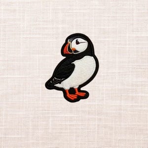 May include: Embroidered patch of a puffin bird. The patch features a black and white body with a red and orange beak and feet. The bird is outlined in black and has a white background.