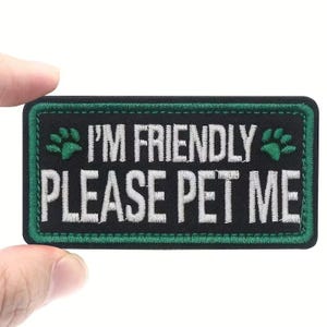 May include: A rectangular black patch with green stitching and border. The patch features the text "I'M FRIENDLY PLEASE PET ME" in white, with green paw prints on either side of "I'M FRIENDLY".