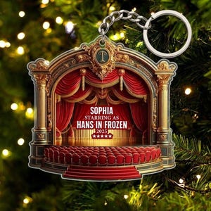 May include: A theater-themed Christmas ornament with a stage design. The ornament features red curtains, gold columns, and a seating area. Text on the ornament reads "Sophia starring as Hans in Frozen 2025". A silver chain and ring are attached for hanging.