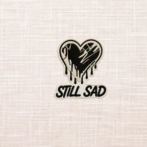 May include: A black and white patch featuring a heart design with dripping details and the text "STILL SAD" in a bold font. The patch is set against a textured, off-white background.