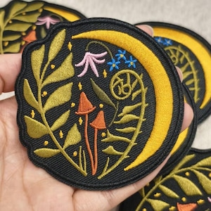 May include: Embroidered patch with a black background featuring a yellow crescent moon, orange mushrooms, and various botanical elements in gold, pink, and blue. The patch has a black border.