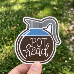 May include: A sticker featuring a coffee pot design with the words "POT head" in a cursive font. The coffee pot has a brown body, blue liquid, and a gray lid and handle. The sticker is white-bordered.