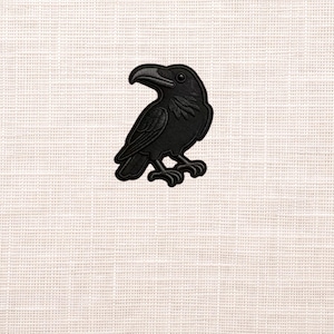 May include: A black raven patch with detailed embroidery. The bird is depicted in a side profile, perched on two feet. The patch has a textured appearance and is set against a neutral, off-white background.