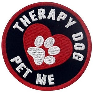 May include: Round patch with a black background and a red border. The patch features a red heart with a white paw print in the center. The words "THERAPY DOG PET ME" are embroidered in white around the heart.
