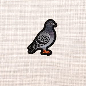 May include: Embroidered patch featuring a stylized pigeon with gray and black feathers, orange feet, and a black outline. The patch is set against a textured, off-white background, suitable for application on clothing or accessories.