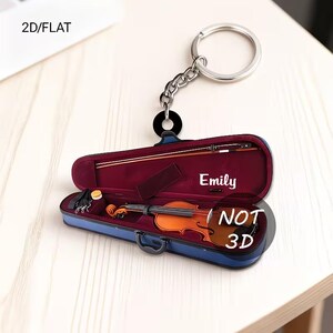 May include: A 2D flat keychain featuring a miniature violin inside a blue case with a red interior. The keychain includes a bow and the name "Emily". The text "NOT 3D" is also visible.