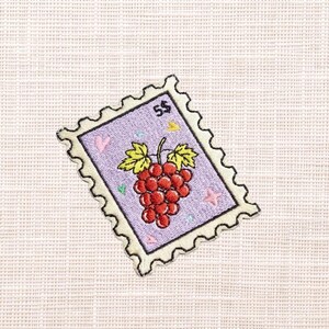 May include: Embroidered patch in the shape of a postage stamp. The stamp features a cluster of red grapes with green leaves, a pink heart, and a yellow star on a lavender background. The stamp is edged with a scalloped border and the number "5$" is in the upper right corner.