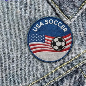 May include: Embroidered patch featuring the text "USA SOCCER" in white on a blue background. The patch includes the American flag and a soccer ball. The patch is round and sewn onto denim.