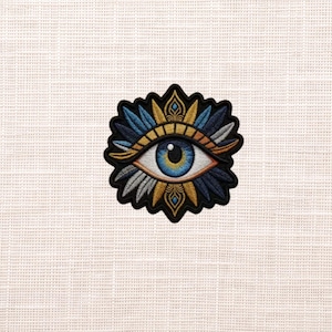 May include: Embroidered patch featuring a stylized eye with blue, gold, and white feather-like details. The eye has a blue iris and a gold and orange eyelid. The patch is outlined in black and is likely for clothing or accessories.