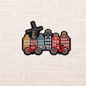 May include: A colorful Dutch-themed design featuring a black windmill, several buildings in various colors, and two red bicycles. The design is outlined in black and set against a neutral background.