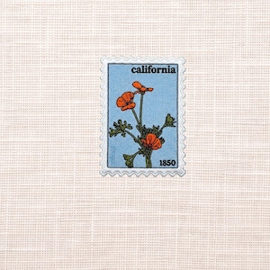 May include: A rectangular embroidered patch shaped like a postage stamp with a light blue background. It features orange California poppy flowers with green stems and leaves. The text "california" is at the top and "1850" is at the bottom.