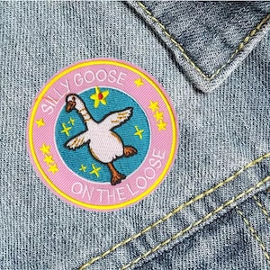 May include: Embroidered patch with a pink and yellow border, featuring a cartoon goose with outstretched wings. The patch reads "SILLY GOOSE ON THE LOOSE" in a circular design, set against a denim background.