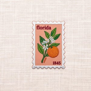 May include: A rectangular embroidered patch shaped like a postage stamp with a peach-colored background. It features a green branch with white blossoms and a single orange fruit. The text reads Florida at the top and 1845 at the bottom in red.